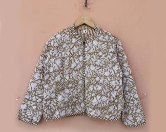 May include: A quilted jacket with a floral pattern in white and olive green. The jacket has two front pockets and a striped collar. The edges are trimmed with a dark brown rope-like material. The jacket is hanging on a wooden hanger.