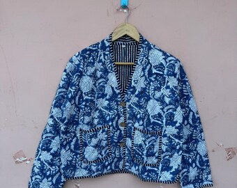 May include: A blue and white floral print jacket with a V-neck, two front pockets, and wooden buttons. The jacket is hanging on a wooden hanger against a pink background. The interior lining has a black and white striped pattern.