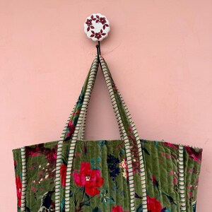 May include: A green quilted tote bag with a floral pattern of red flowers and green leaves. The bag has striped handles and trim. A decorative hook with a floral design holds the bag against a pink wall.