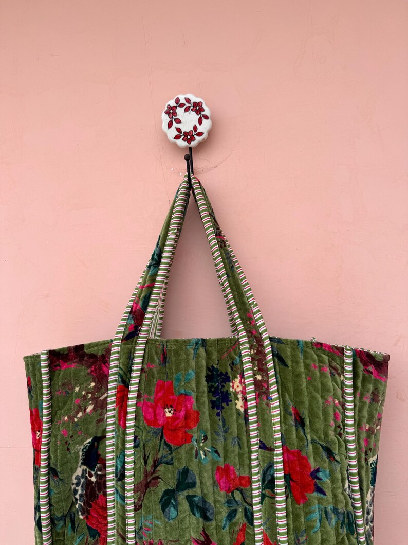 May include: A green quilted tote bag with a floral pattern of red flowers and green leaves. The bag has striped handles and trim. A decorative hook with a floral design holds the bag against a pink wall.