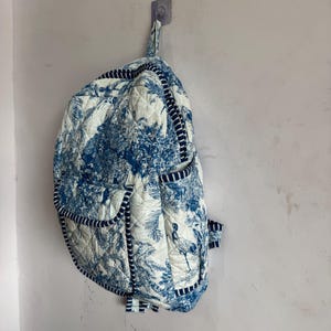 Peut inclure: Un sac &agrave; dos matelass&eacute; avec un motif de toile de Jouy bleu et blanc repr&eacute;sentant des arbres et des oiseaux. Le sac &agrave; dos a une bordure et des bretelles bleu marine, et est accroch&eacute; &agrave; un mur. Le sac a plusieurs poches et une poign&eacute;e sup&eacute;rieure.