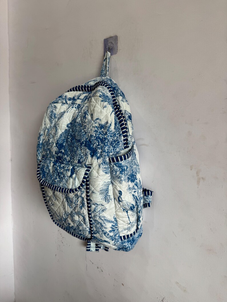 Peut inclure: Un sac &agrave; dos matelass&eacute; avec un motif de toile de Jouy bleu et blanc repr&eacute;sentant des arbres et des oiseaux. Le sac &agrave; dos a une bordure et des bretelles bleu marine, et est accroch&eacute; &agrave; un mur. Le sac a plusieurs poches et une poign&eacute;e sup&eacute;rieure.