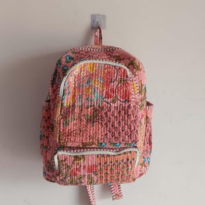 以下が含まれることがあります： ピンク色のキルトバックパック。花柄と白いトリムが特徴です。バックパックには上部のハンドル、フロントポケット、サイドポケットがあります。生地は質感があり、デザインにはさまざまな色が使用されています。