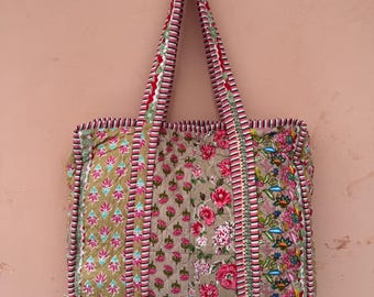 Könnte beinhalten: Eine gesteppte Tragetasche mit floralen Mustern in Rosa, Grün und Beige. Die Tasche hat ein Patchwork-Design mit vertikalen Streifen und roten und weißen Zierleisten. Die Griffe sind ebenfalls gemustert und rot und weiß eingefasst.