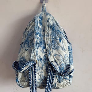 Peut inclure: Un sac &agrave; dos matelass&eacute; avec un motif de toile de Jouy bleu et blanc repr&eacute;sentant des arbres et des oiseaux. Le sac &agrave; dos a des bretelles et des poches lat&eacute;rales ray&eacute;es bleues et blanches. Le sac est accroch&eacute; &agrave; un mur.