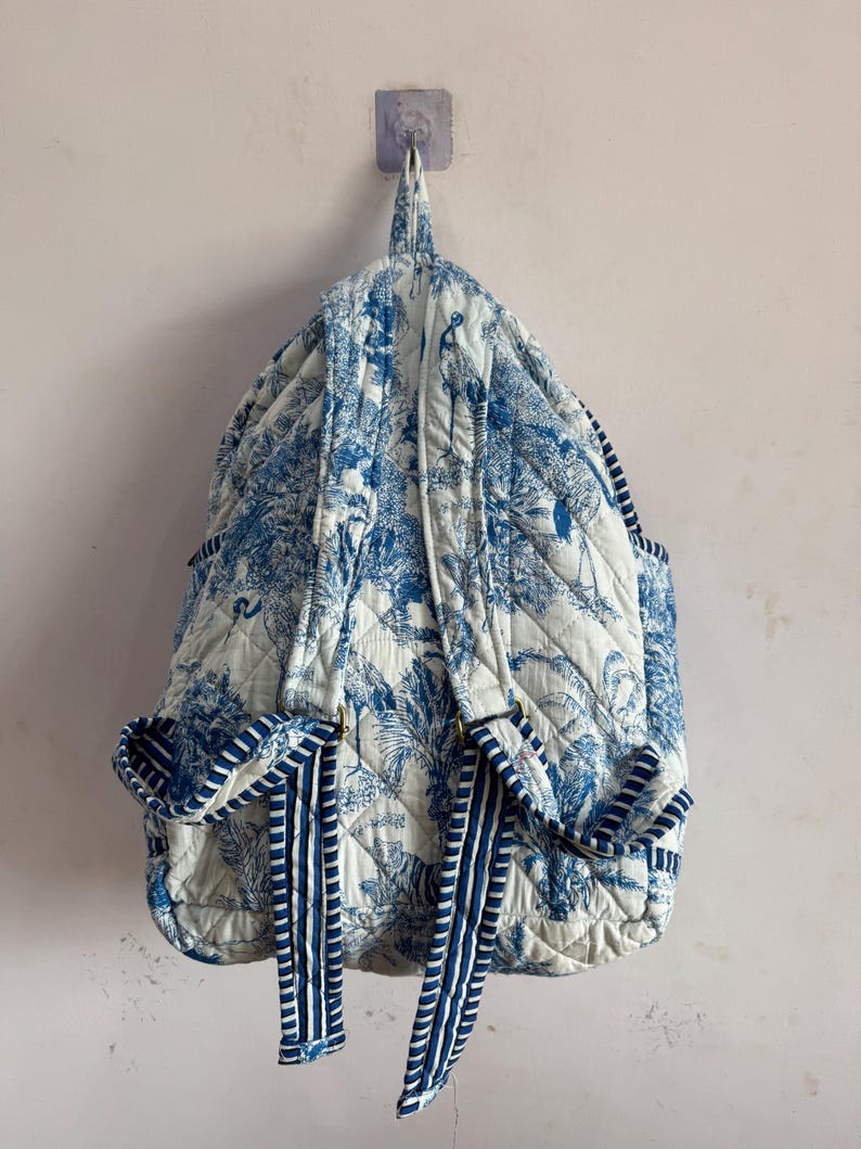 Peut inclure: Un sac &agrave; dos matelass&eacute; avec un motif de toile de Jouy bleu et blanc repr&eacute;sentant des arbres et des oiseaux. Le sac &agrave; dos a des bretelles et des poches lat&eacute;rales ray&eacute;es bleues et blanches. Le sac est accroch&eacute; &agrave; un mur.