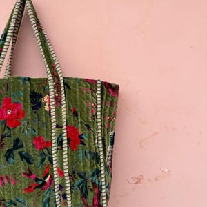 May include: A green quilted tote bag with a floral pattern featuring red flowers and birds. The bag has striped handles and trim. The background is a solid pink color.