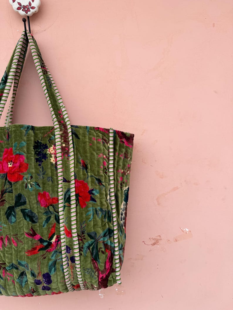May include: A green quilted tote bag with a floral pattern featuring red flowers and birds. The bag has striped handles and trim. The background is a solid pink color.