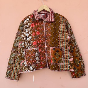 May include: A quilted jacket with a patchwork design featuring floral and geometric patterns in shades of brown, red, and white. The jacket has a striped collar and two front pockets. The jacket is hanging on a wooden hanger.