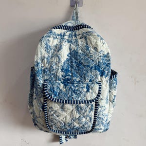 May include: A quilted backpack with a blue and white toile pattern. The backpack has a flap pocket, side pockets, and a top handle. The edges are trimmed with a blue and white striped fabric. The backpack is hanging on a wall.