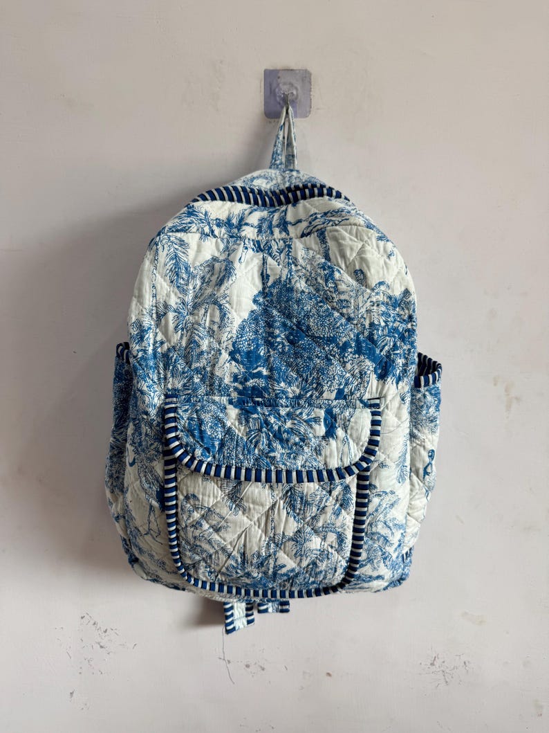 Peut inclure: Sac &agrave; dos matelass&eacute; avec un motif de toile de Jouy bleu et blanc. Il comporte une poche &agrave; rabat, des poches lat&eacute;rales et une poign&eacute;e sup&eacute;rieure. Les bords sont bord&eacute;s d'un tissu ray&eacute; bleu et blanc. Le sac est accroch&eacute; au mur.