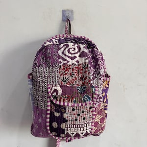 Puede incluir: Una mochila de patchwork morada, con varios cuadrados de tela estampada. La mochila tiene un bolsillo frontal, bolsillos laterales y un asa superior. El diseño incluye patrones florales y geométricos, con una textura acolchada.