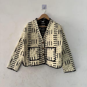 Peut inclure: Veste matelassée crème avec un motif géométrique noir. La veste a un col en V, deux poches avant et est boutonnée. Elle est suspendue à un cintre en bois sur un mur blanc.
