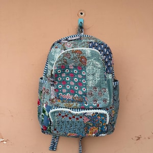 Puede incluir: Mochila acolchada con diseño de patchwork en tonos azules, verdes y rosas. La mochila presenta estampados florales y ribete blanco. Tiene un asa superior y correas ajustables.