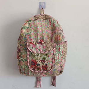 Può includere: Uno zaino trapuntato con motivo floreale. Il design presenta fiori rossi, rosa e verdi su sfondo beige. Ha due tasche laterali e una tasca frontale, tutte bordate con tessuto a righe rosa e bianco.