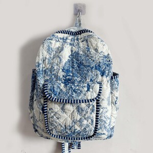 Peut inclure: Un sac &agrave; dos matelass&eacute; blanc et bleu, orn&eacute; d'un motif de toile de Jouy repr&eacute;sentant un paysage. Le sac &agrave; dos est dot&eacute; d'une poche avant, de poches lat&eacute;rales et d'une bordure ray&eacute;e. Il est accroch&eacute; au mur.