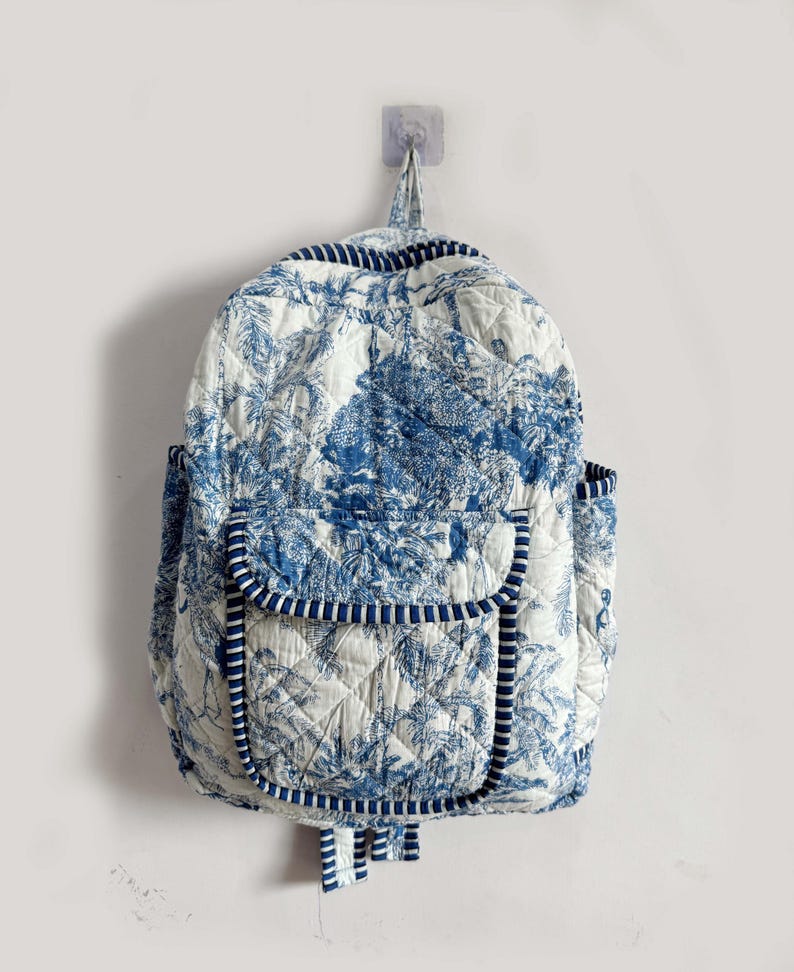 Peut inclure: Un sac &agrave; dos matelass&eacute; blanc et bleu, orn&eacute; d'un motif de toile de Jouy repr&eacute;sentant un paysage. Le sac &agrave; dos est dot&eacute; d'une poche avant, de poches lat&eacute;rales et d'une bordure ray&eacute;e. Il est accroch&eacute; au mur.