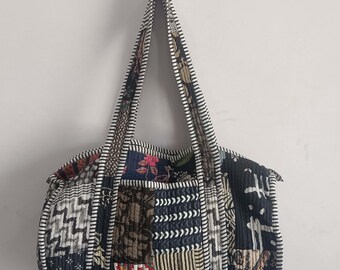 Könnte beinhalten: Eine Patchwork-Tragetasche mit einer Vielzahl von gemusterten Stoffquadraten in Blau-, Braun- und Weißtönen. Die Tasche hat lange Griffe und eine gesteppte Textur. Die Tasche hängt an einer weißen Wand.