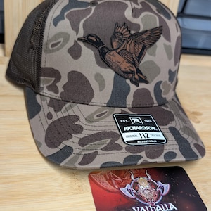 Puede incluir: Gorra de camionero de camuflaje con parte trasera de malla marrón y un parche de pato. La gorra tiene una visera curva y una correa ajustable. El parche presenta una ilustración detallada de un pato en vuelo. La gorra tiene el texto "RICHARDSON 112 TRUCKER" en una etiqueta.