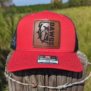 May include: Red and black trucker hat with a brown leather patch that says "DAWGS" and features a bulldog head. The hat is adjustable and has the Richardson 112 logo.