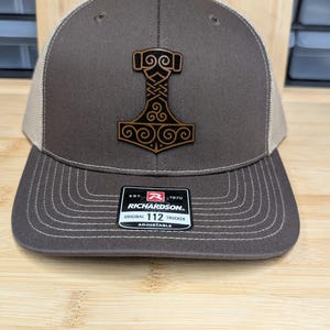 May include: A brown and tan trucker hat with a leather-like patch featuring a stylized hammer design. The hat has a curved brim and a mesh back. The Richardson 112 label is visible on the front.