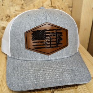 Può includere: Un cappello da camionista grigio e bianco con una toppa in pelle marrone che presenta una bandiera americana e una silhouette di un lineman che lavora su una linea elettrica.