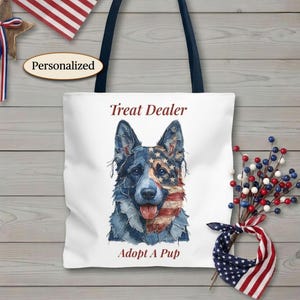 Puede incluir: Un bolso tote blanco con asas azul oscuro que presenta un gráfico de un pastor alemán con un patrón de mezclilla y bandera estadounidense. El texto Treat Dealer está impreso sobre el perro y Adopt A Pup debajo en fuente roja.