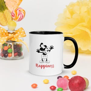 Może przedstawiać: Biały ceramiczny kubek z czarnym wnętrzem i uchwytem. Kubek przedstawia czarno-białą ilustrację myszy z kresk&oacute;wki ze słowem "Happiness" na czerwono. Kubek otoczony jest kolorowymi cukierkami i ż&oacute;łtym kwiatem.