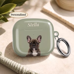 Pode incluir: Uma capa protetora verde-sálvia para auriculares sem fios com o retrato impresso de um bulldog francês tigrado. O nome Stella está impresso em branco por cima do cão. Um anel mosquetão de metal prateado está preso na lateral.