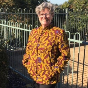 May include: A woman wearing a yellow and brown patterned shirt with a mandarin collar and long sleeves. The shirt has a floral design with red, blue, and green accents.
