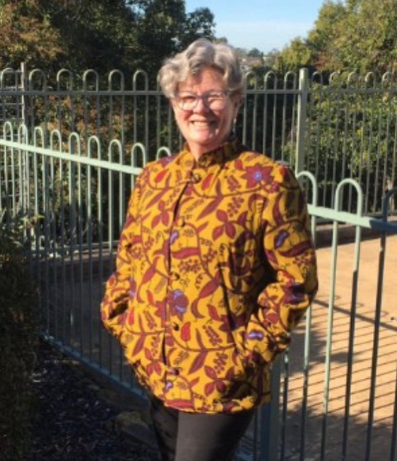 May include: A woman wearing a yellow and brown patterned shirt with a mandarin collar and long sleeves. The shirt has a floral design with red, blue, and green accents.