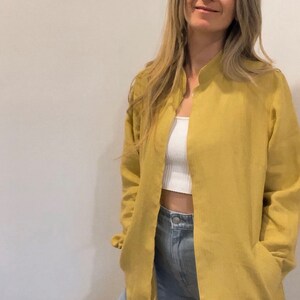 May include: A long-sleeved, mustard yellow linen shirt with a mandarin collar and a single button closure at the neck. The shirt is open in the front, revealing a white tank top underneath. The model is wearing blue jeans.
