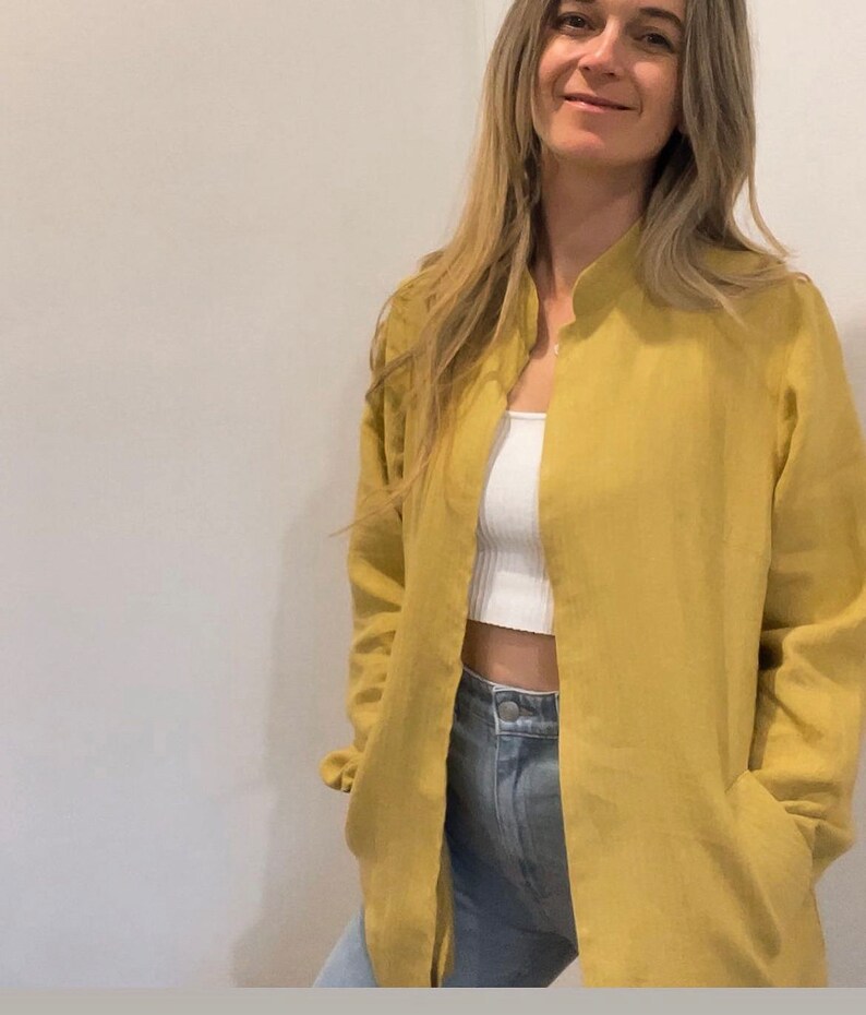 May include: A long-sleeved, mustard yellow linen shirt with a mandarin collar and a single button closure at the neck. The shirt is open in the front, revealing a white tank top underneath. The model is wearing blue jeans.