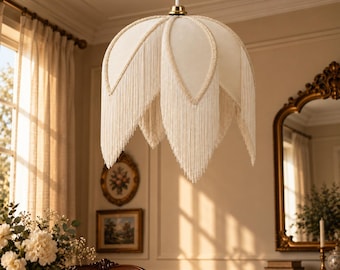 May include: A cream-colored pendant light fixture with a petal-shaped shade featuring long, hanging fringe trim along the edges. The lamp is suspended by a white cord and includes a gold-toned metal cap at the top where the shade connects.