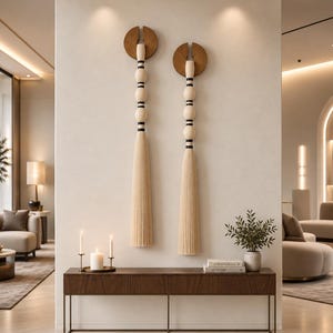 May include: Two vertical wall hangings featuring a circular wooden top, a series of stacked light-coloured wooden spheres separated by thin black rings, and a long, pleated, light-coloured fabric tassel hanging from the bottom of each piece.