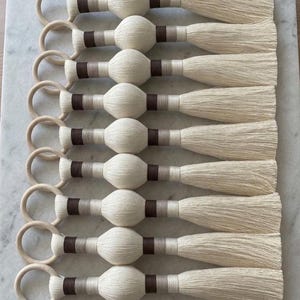 May include: Nine decorative tassels arranged in a vertical row. Each tassel features a light wooden ring at the top, followed by a central sphere wrapped in cream-colored thread, accented with horizontal bands of dark brown and light gray thread.