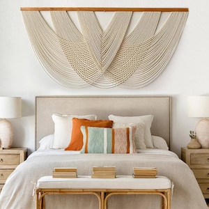 May include: A bedroom interior with a bed, beige headboard, and cream-colored macrame wall hanging. The bed is dressed with pillows and a light-colored duvet. Two wooden bedside tables with lamps are on either side of the bed, and a bench is at the foot.