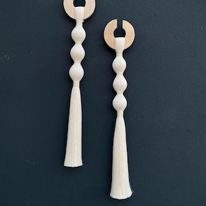 Op de afbeelding: Twee witte kwast wanddecoraties met houten ringen aan de bovenkant. Elke kwast heeft vier witte kralen.