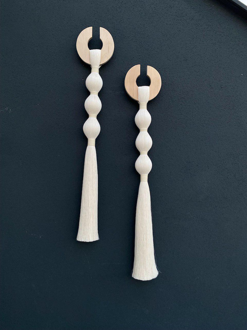 Op de afbeelding: Twee witte kwast wanddecoraties met houten ringen aan de bovenkant. Elke kwast heeft vier witte kralen.