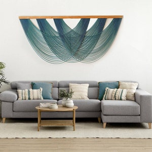 May include: A gray sectional sofa with a wooden coffee table in front of it. The wall behind the sofa features a large, teal and blue macrame wall hanging.