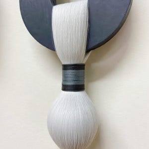 May include: A modern wall hanging featuring a dark gray circular wooden element with a white, textured, brush-like design. The brush is bound with black and gray thread. The artwork is a minimalist, abstract design.
