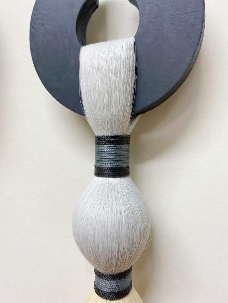 May include: A modern wall hanging featuring a dark gray circular wooden element with a white, textured, brush-like design. The brush is bound with black and gray thread. The artwork is a minimalist, abstract design.
