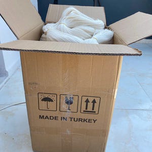 Op de afbeelding: Een kartonnen doos gevuld met wit garen, met de woorden "MADE IN TURKEY" erop gedrukt. De doos heeft drie symbolen erop gedrukt: een paraplu, een glas en een pijl die omhoog wijst.