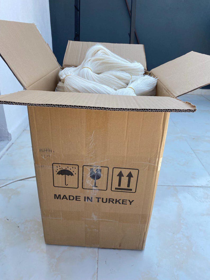 Op de afbeelding: Een kartonnen doos gevuld met wit garen, met de woorden "MADE IN TURKEY" erop gedrukt. De doos heeft drie symbolen erop gedrukt: een paraplu, een glas en een pijl die omhoog wijst.