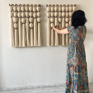 Op de afbeelding: Twee beige macram&eacute; wandhangers met houten latten en kwastjes. De hangers zijn gemaakt van dik garen en hebben een geweven textuur.