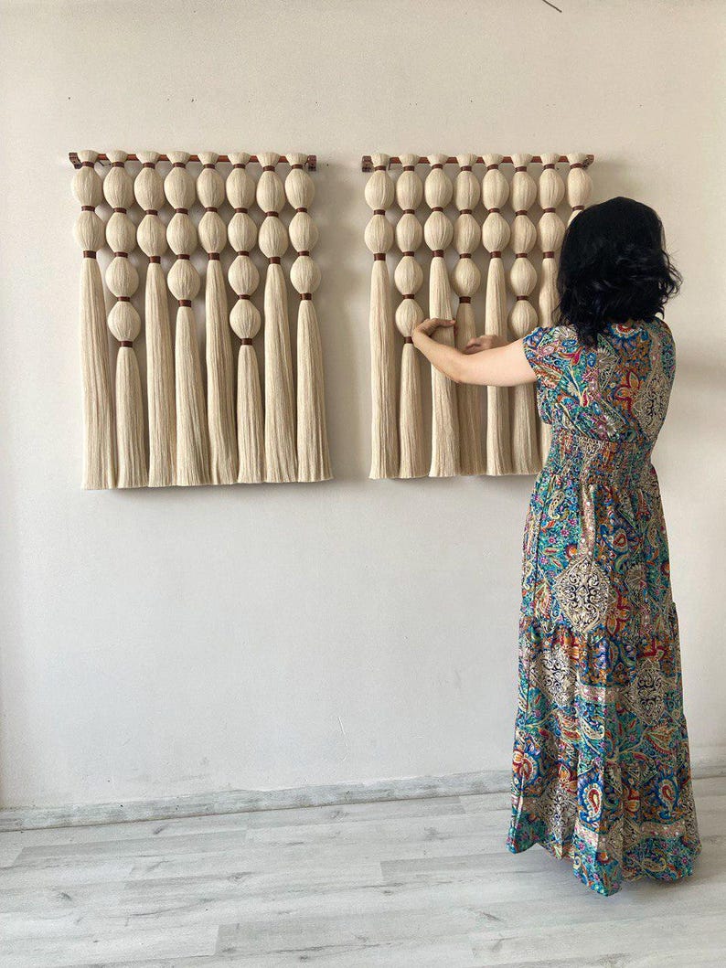 Op de afbeelding: Twee beige macram&eacute; wandhangers met houten latten en kwastjes. De hangers zijn gemaakt van dik garen en hebben een geweven textuur.