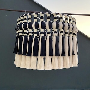 Op de afbeelding: Een zwart-witte macram&eacute; hanglamp met kwastjes. De lamp is gemaakt van katoenen koord en heeft een ronde vorm.