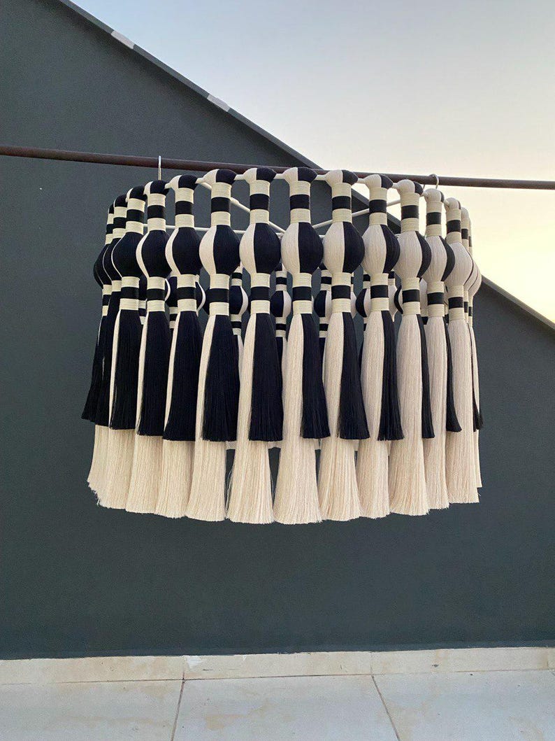 Op de afbeelding: Een zwart-witte macram&eacute; hanglamp met kwastjes. De lamp is gemaakt van katoenen koord en heeft een ronde vorm.