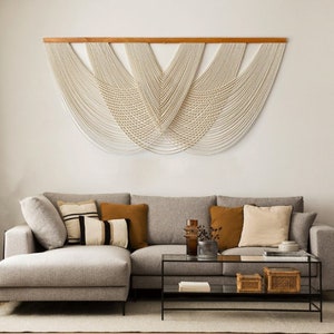 May include: A light grey sectional sofa with a chaise lounge, featuring a variety of throw pillows in beige, brown, and black and white stripes. The sofa is positioned in front of a white wall with a large macrame wall hanging in cream.