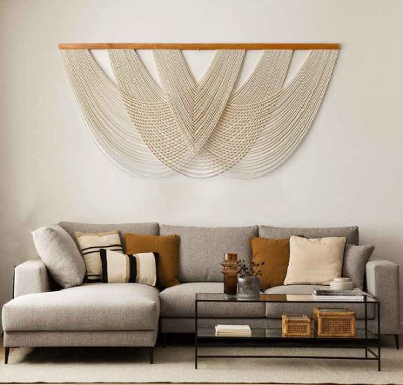 May include: A light grey sectional sofa with a chaise lounge, featuring a variety of throw pillows in beige, brown, and black and white stripes. The sofa is positioned in front of a white wall with a large macrame wall hanging in cream.